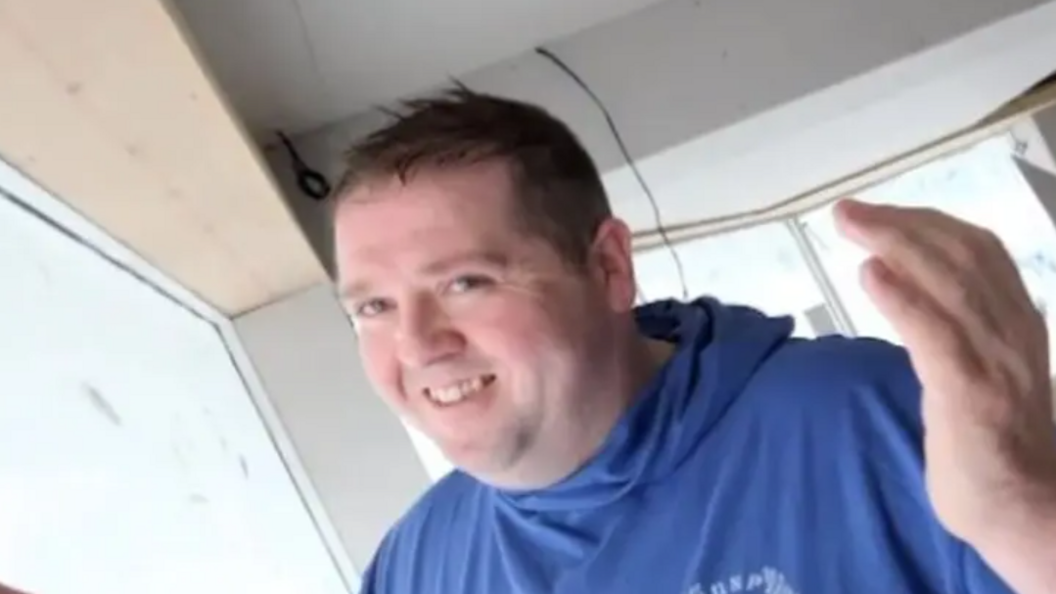 A man with short brown hair wearing a blue hoody smiling