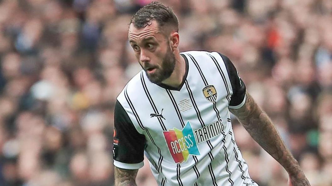 Connell Rawlinson wearing a white Notts County shirt with a black pinstripe, black shorts and black socks controls the ball while playing against Chesterfield at Wembley