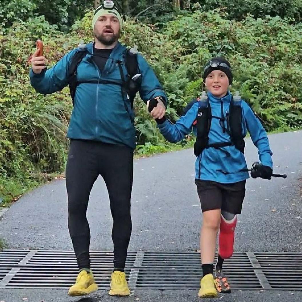 Albie-Junior and his dad Daniel Thomas completing the Three Peaks challenge. The pair are both wearing beanie hats, with flash lights attached to them. They are both wearing blue long sleeve tops, matching brought yellow trainers. The pair are hand in hand as they walk towards the camera.