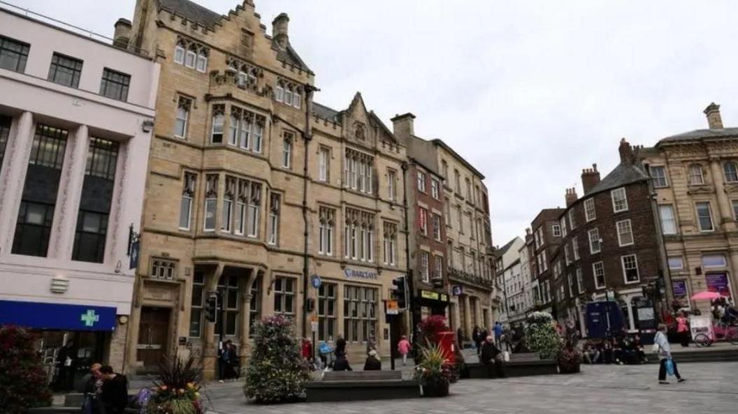 Durham city centre on an overcast day. There is a large brown-bricked branch of Barclays bank to the left, some seats and shrubs and a few shoppers walking past. 