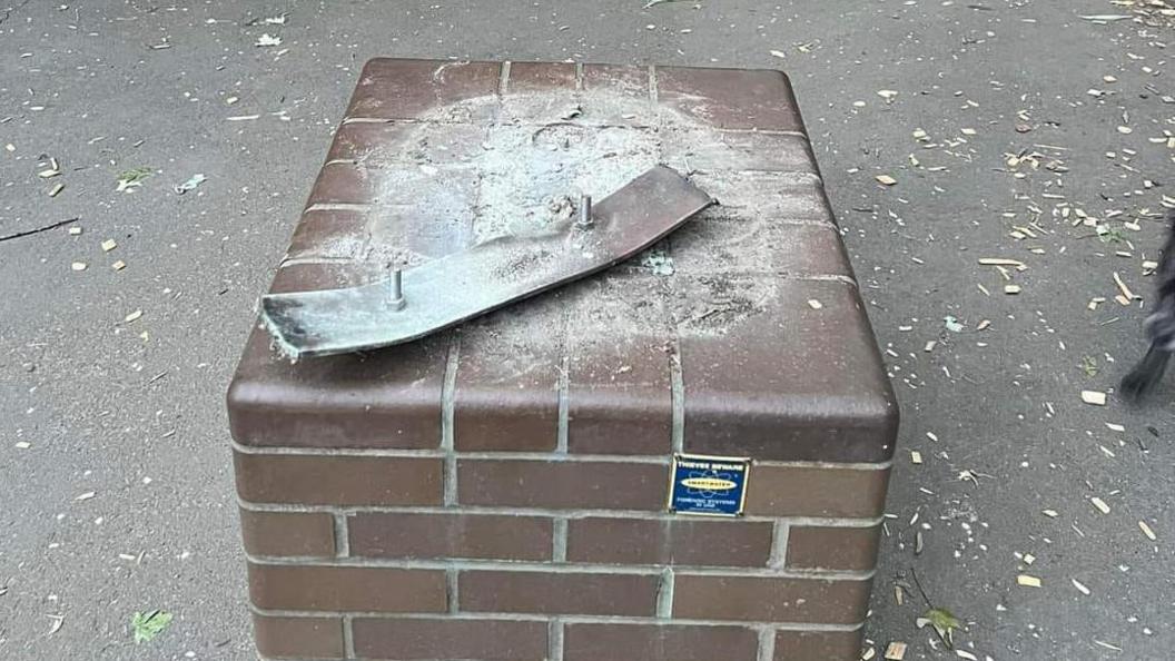 The empty plinth where the statue once stood. It is a square brown-brick platform a foot or so high. The statue has gone, there is just a metal plate with screws on which once held it, which appears to have been partly wrenched from the plinth.