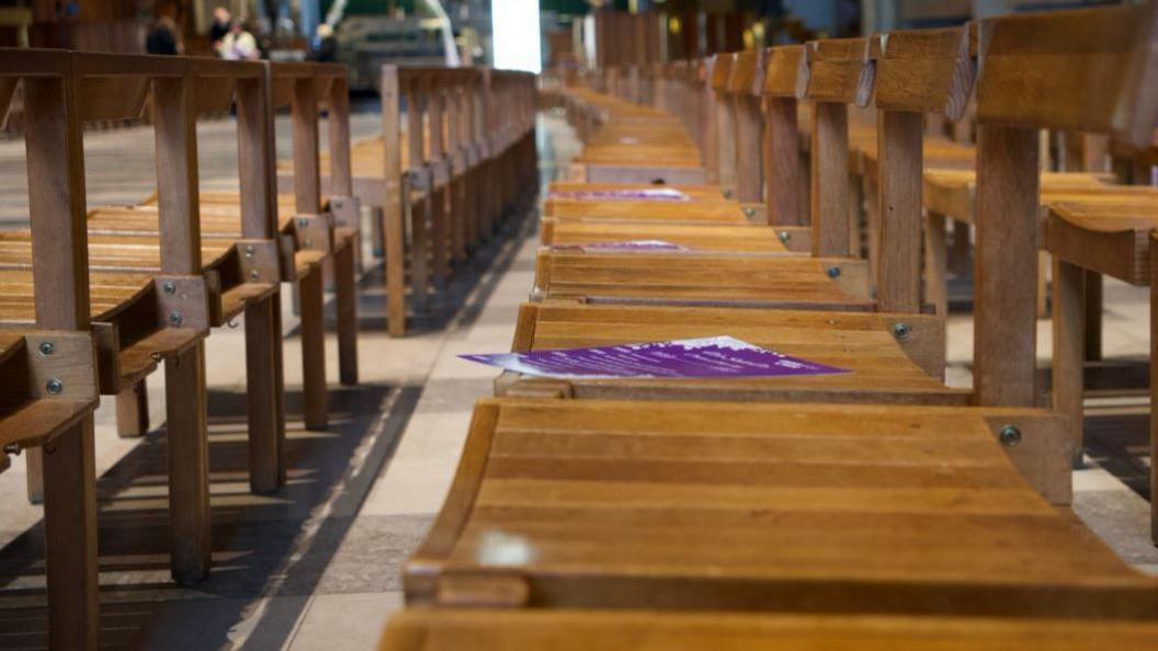 A close-up of row of wooden chairs in a cathedral, with purple leaflets on every other seat. The row is otherwise empty.