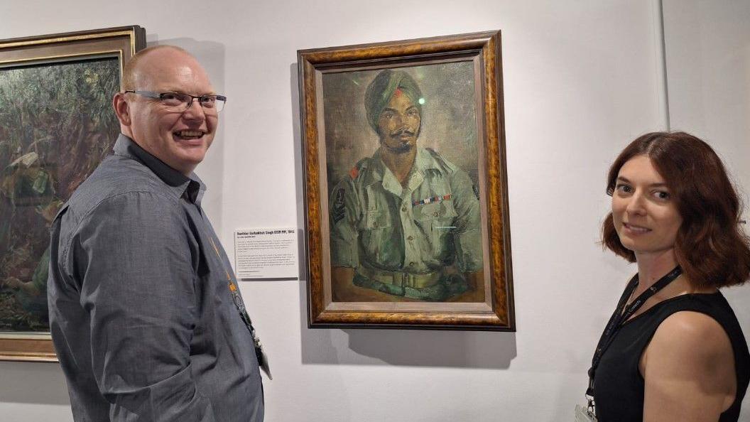 John Wood and Katie Ackrill stand next to Leslie Cole's Portrait Of Indian Army Sergeant. They are looking at the camera, smiling.