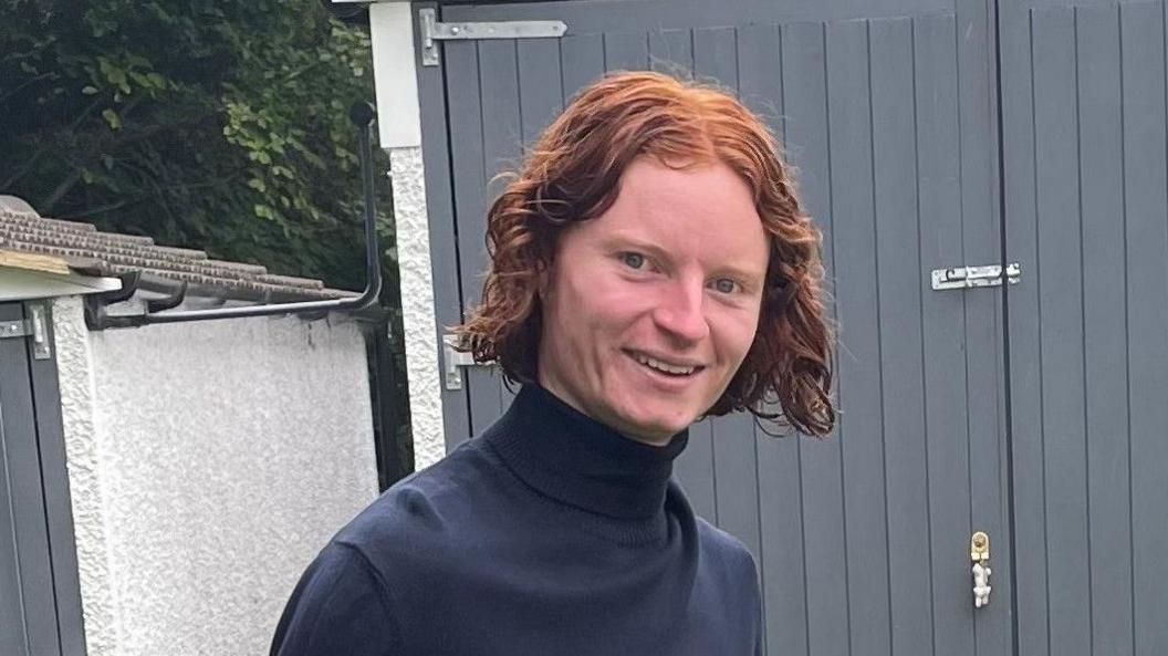 George Gilbert is wearing a navy turtle neck jumper in front of a shed in a garden. He is smiling.