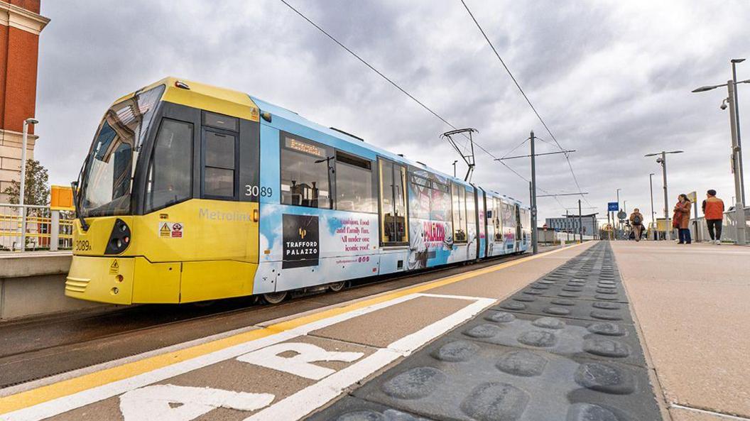A tram at the stop outside the Trafford Centre. The driver's cab is yellow and the rest of the tram has been decorated with Trafford Palazzo branding