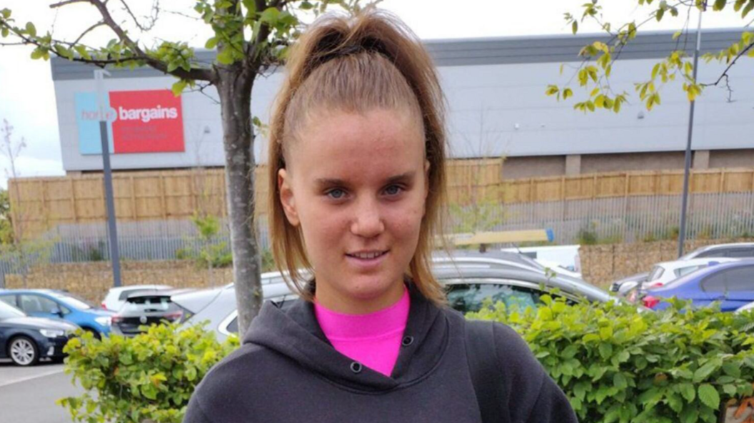 Holly Newton, photographed in a car park with trees and vehicles behind her, has long dark hair tied up in a pony tail and is wearing a pink shirt under grey hoodie. She is looking directly at the camera