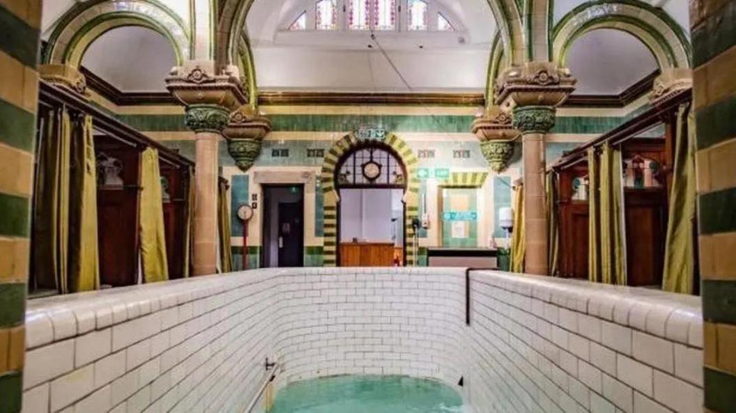 A highly decorated room with green and mustard tiles and ornate columns. There is a deep plunge pool in the foreground with white tiles. At the sides there are booths separated by wooden panels with decorations at the top and green privacy curtains.
