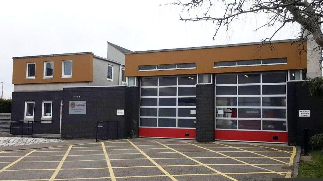 The fire station is a square box. It has roller shutters at the front with fire engines behind. It is a brown and black building.