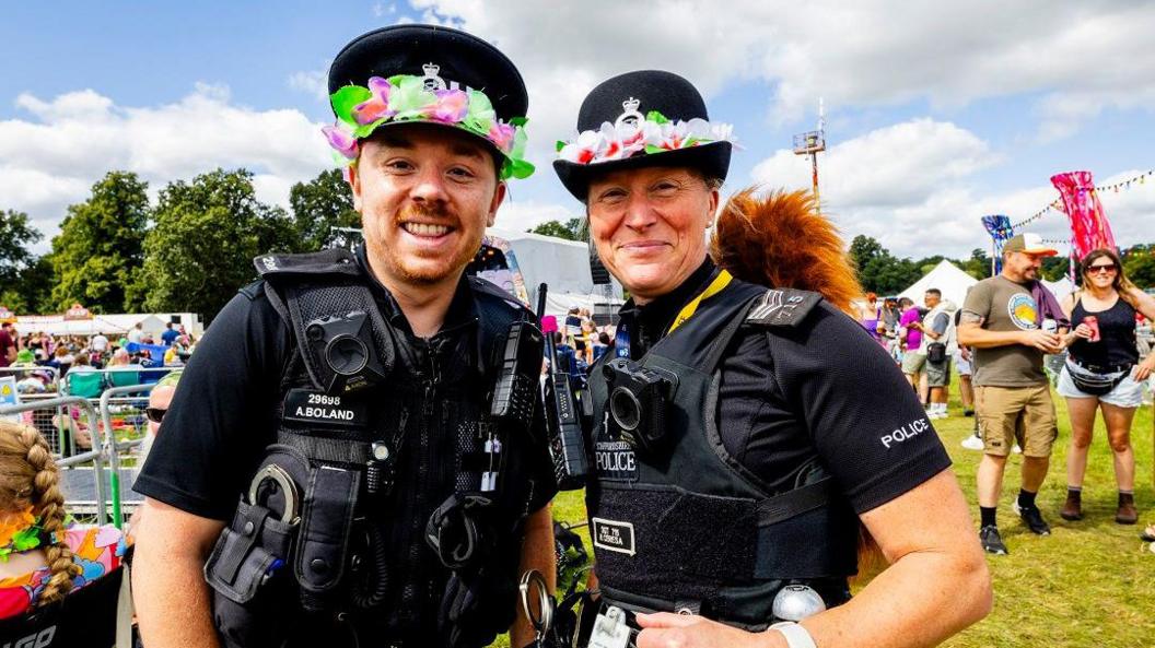 Two police officers in uniform, both with flowery headbands around their headwear, smile at the camera. One is male, one if female and behind them are festival-goers