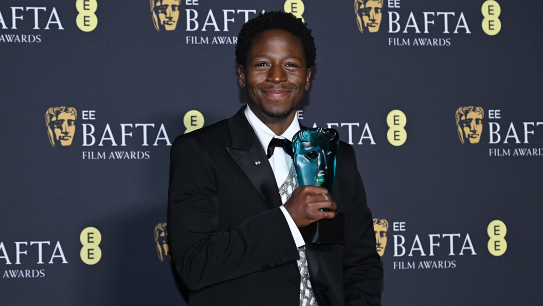 David Jonsson stands in front of a hoarding with "EE Bafta Film Awards" printed on it. He's wearing a black suit with white shirt and bow tie.