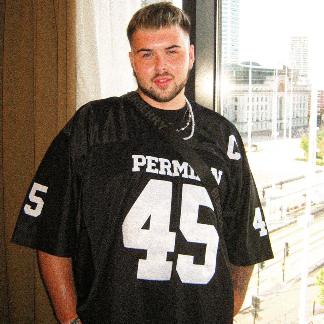 Jak Howells, a man with brown hair and a beard stands in front of a window. He is wearing a black American Football jersey and has tanned arms.
