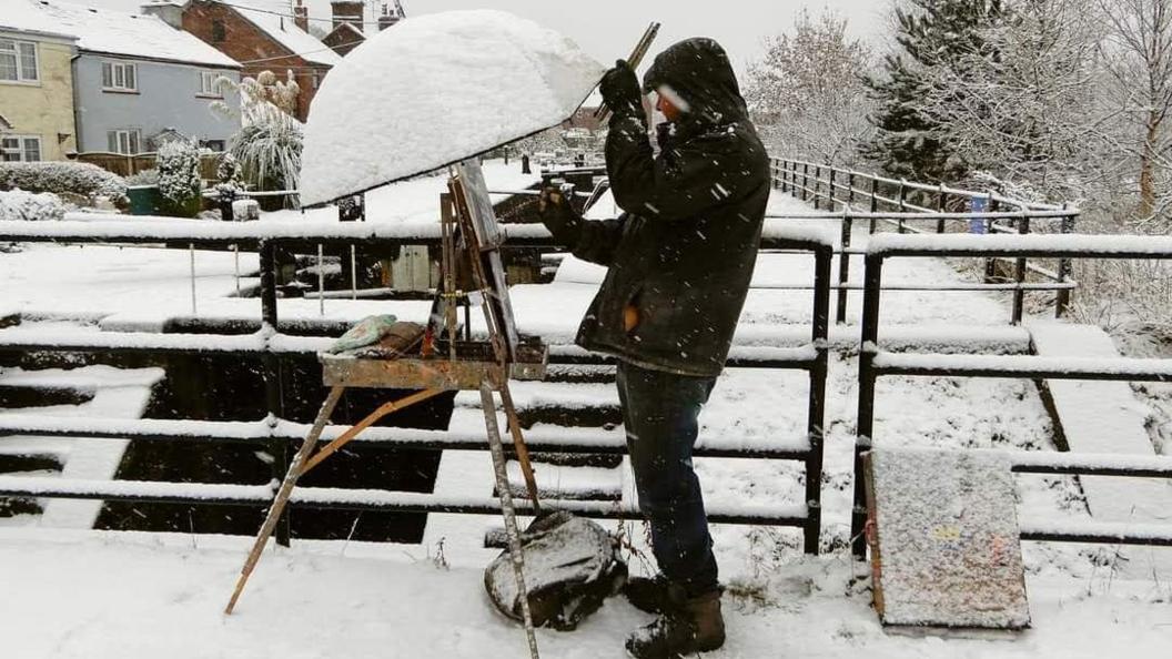 A man paints by a lock in heavy snow. His easel is covered by an umbrella. He is standing by a fence and houses.