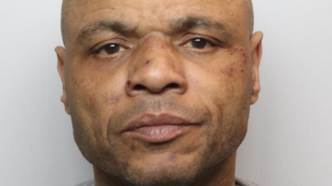 A mugshot of a middle-aged man with a shaved head and a number of sores on his face.