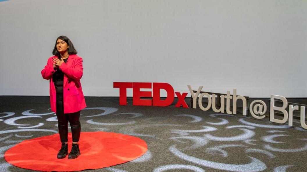 A woman wearing a long pink blazer, black leggings and boots stands on a red circle. The words Tedx Youth Brum can be seen behind her.
