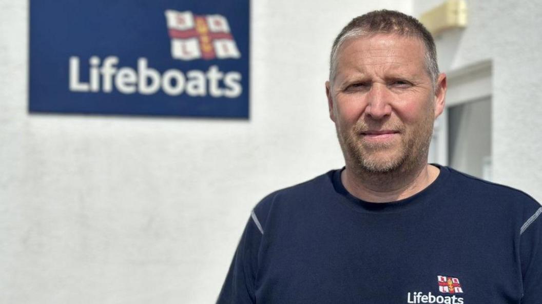 A man with a blue RNLI Lifeboats t shirt is looking directly at the camera. Behind him is a white exterior wall with a blue RNLI Lifeboats sign.
