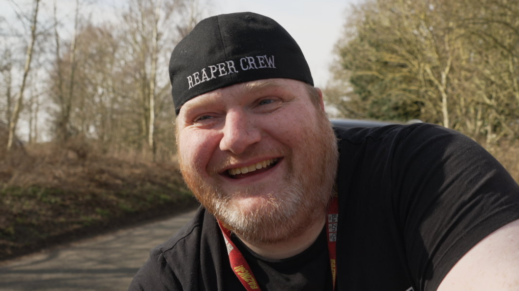 A man smiling while wearing a black tshirt, a basbeball cap on back to front with Reaper Crew in white lettering across it