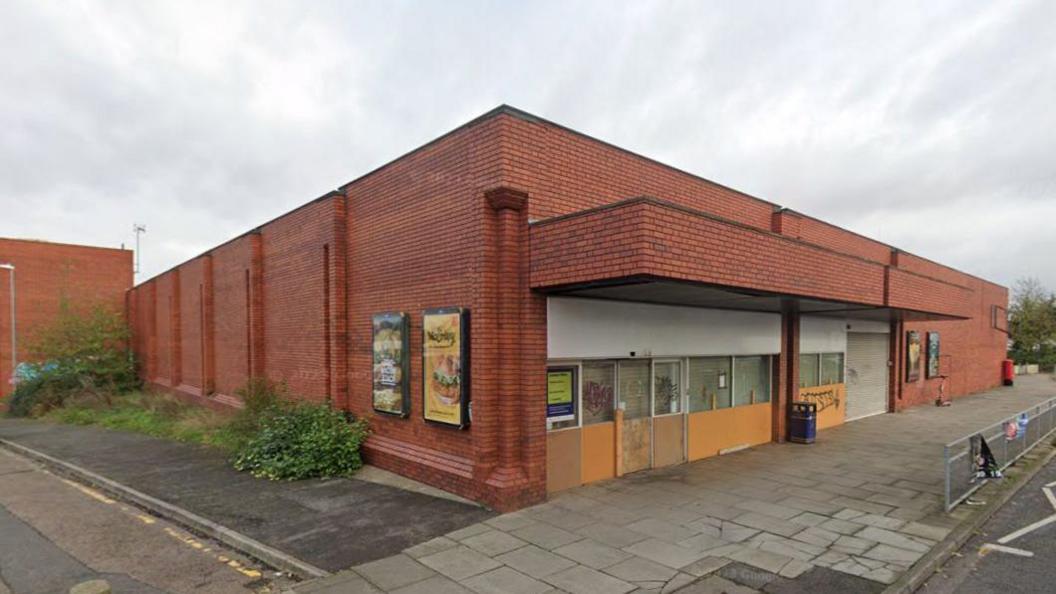 A red brick building with boarded up windows and doors by a tarmac road.