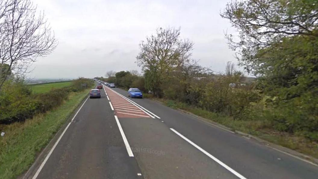 A stretch of dual carriageway with hedgerows and trees on both sides and cars travelling in both directions