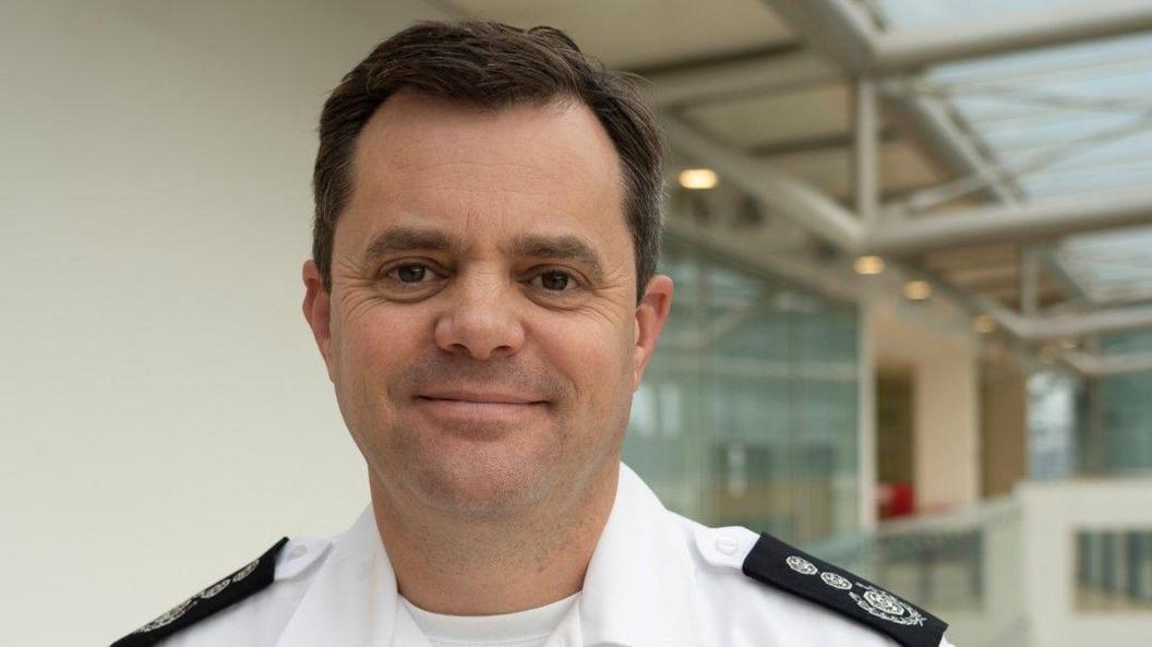 Jon Lacey with dark hair wearing a white shirt with navy fire service epaulettes. he is smiling and looking at the camera. He is stood inside a modern building.