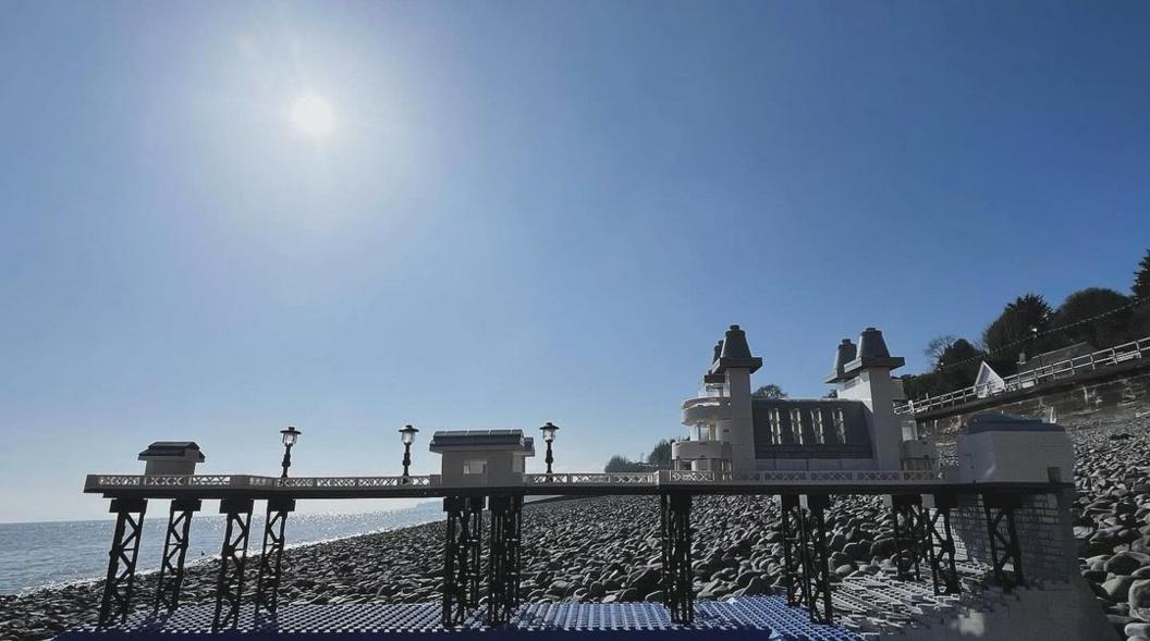 A Lego model of Penarth Pier, sat on the pebbled Penarth beach with the sea in front and on a blue sky and sunshine backdrop