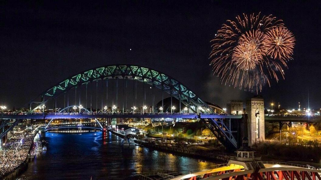 Fireworks exploding in the night sky over the Gateshead quayside, above the Tyne Bridge. In the background, the illuminated Millennium bridge is visible over the river Tyne, as is the Newcastle quayside, packed with crowds.