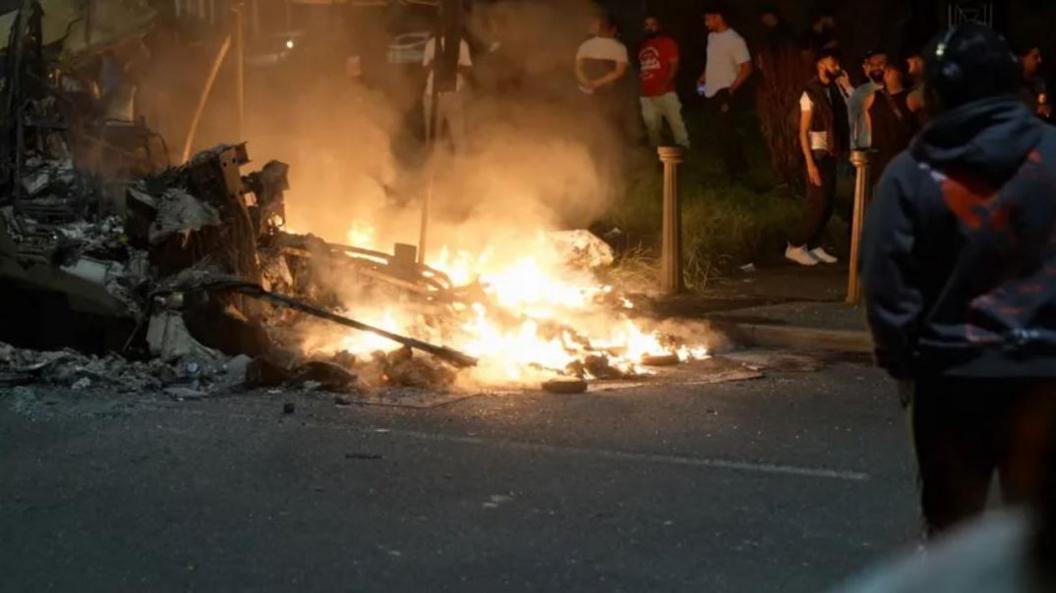 A bus burns in the street as a crowd looks on