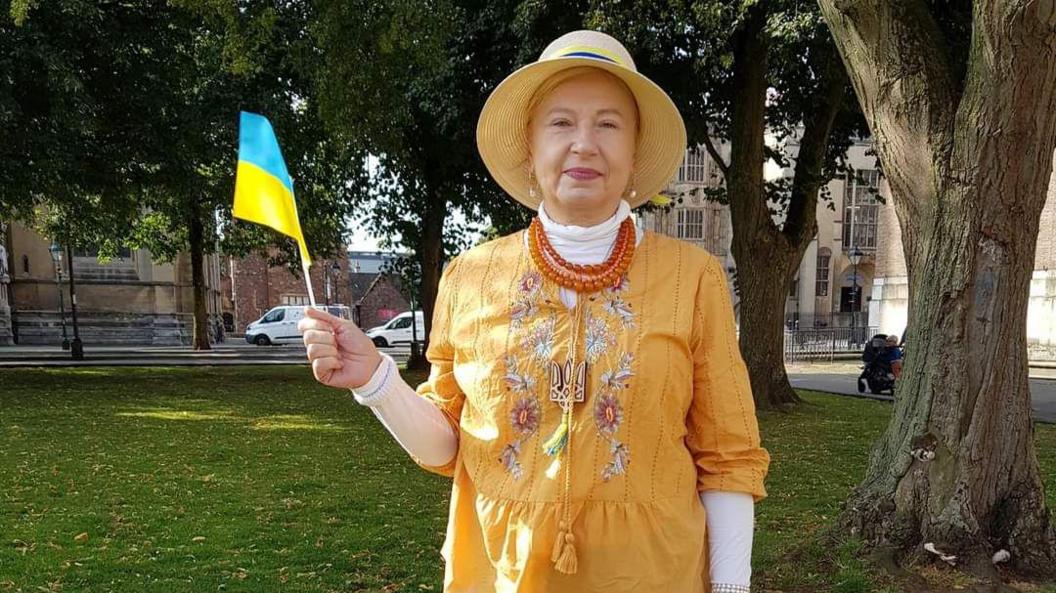 A woman in a mustard outfit and straw hat, waving a mini Ukrainian flag