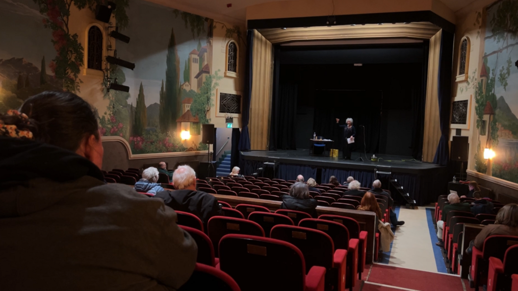 Members of the public sit spread out on the red chairs of the Regal's auditorium. On the stage a woman is addressing the attendees with a microphone.