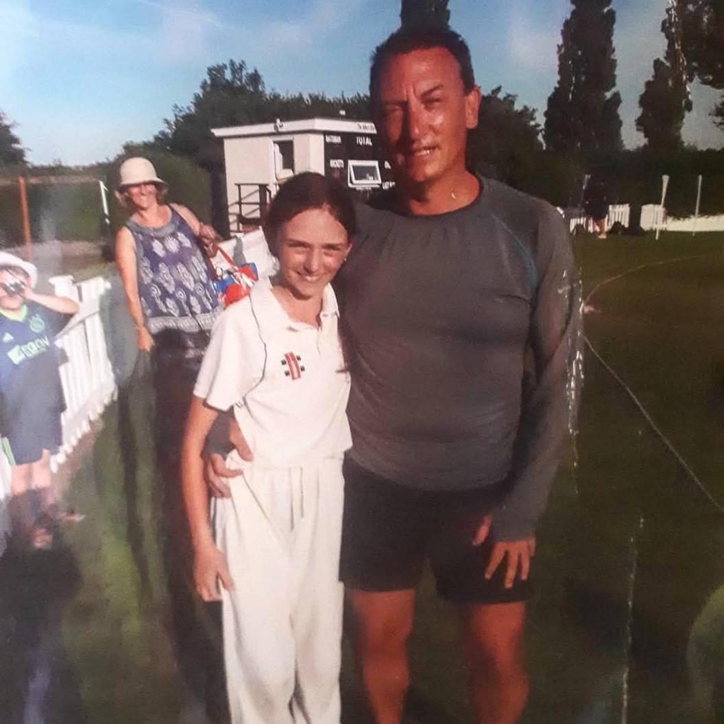 Shortly after representing Wales, Libby is hugged around the waist by her father Michael. Libby is in a white kit with " Cricket Wales / Criced Cymru" logo. A tanned Michael is beaming. 