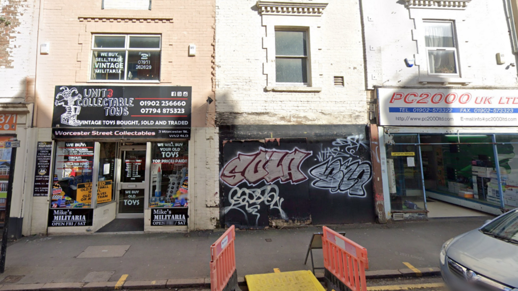 The building sits in between two businesses. The top half of the facade is white brick, with a window and an air vent. The lower half of the building is covered by black boards covered in graffiti that have been put in place. One business on the left looks to be a collectables toy shop and on the right a printing or printer-related store.