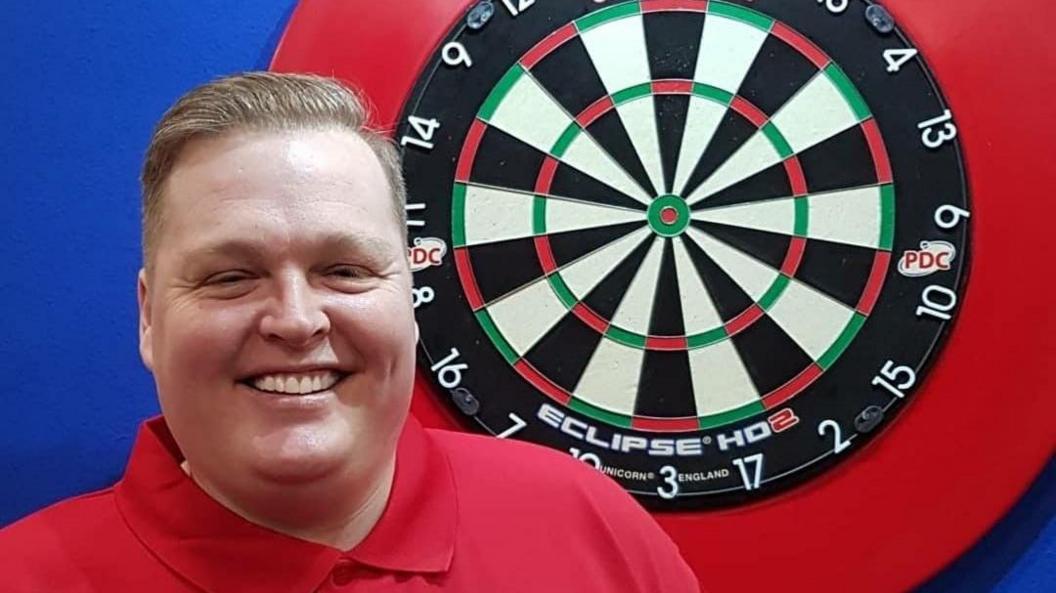 Simon Tate is standing next to a darts board. He is wearing a red top