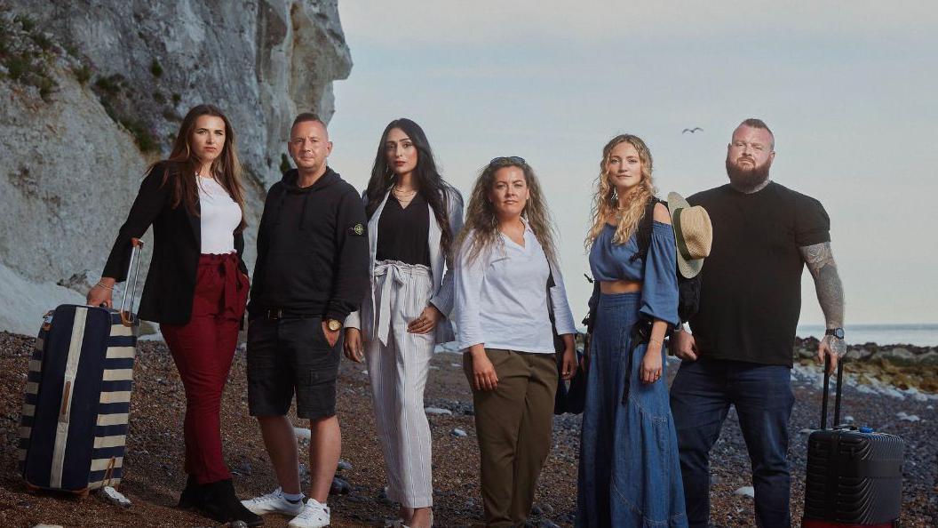 Six people on a beach in Dover - smartly dressed with bags and suitcases. Cliffs on the left, sea on the right, pebble beach with rocks