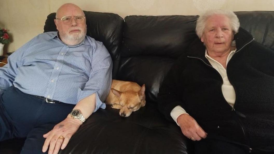 Ian Dunn with shaved head, white beard, blue and white striped shirt with rolled up sleeve and navy blue trousers.
Christine Butterworth with white hair, white coloured sweat shirt and black zip up cardigan  and their little dog in the middle of their black leather sofa.