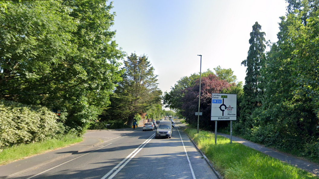 London Road in Guildford - a long, straight road, with a road sign for a roundabout, several cars, trees and grass on either side, a cycle path and a pavement.