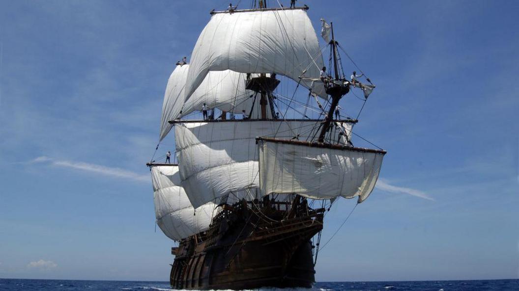 A historic wooden ship with about half a dozen large white sails billowing in the wind. The ship is on the open ocean, with no land visible on the horizon. It is a sunny day, with a clear blue sky above it.