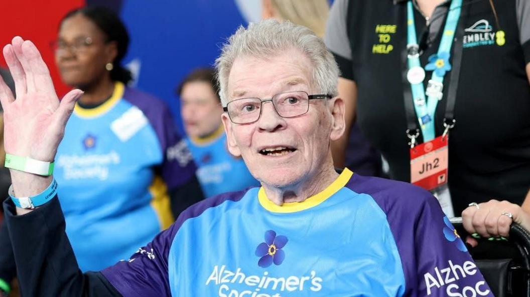 A picture of Adron sitting among the crowd at Wembley, waving at the camera. He had grey hair, is wearing glasses and also a blue and purple football shirt, with a yellow collar, with the Alzheimer's Society logo written on it.