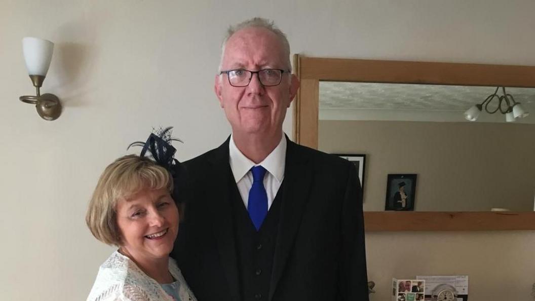 A smiling Jayne Radley (left) wears a black fascinator and pale blue-and-white dress and white patterned scarf. Next to her, her husband Steve wears a black suit, white shirt and dark blue tie.