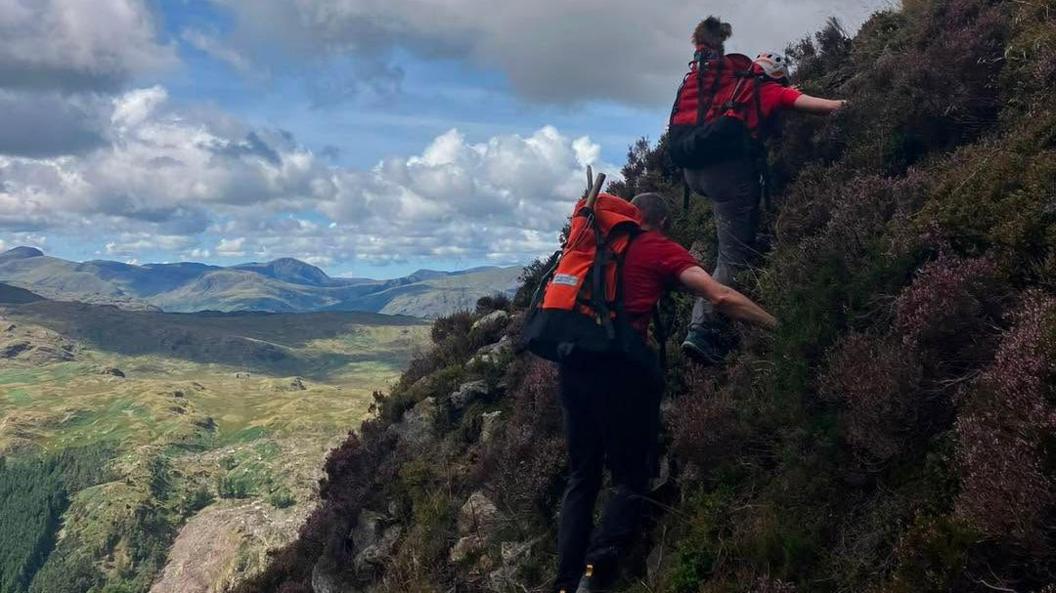 Teddy in the rucksack of a rescuer who is coming down a mountain with another person. There is a dramatic view of the Lake District fells behind them.