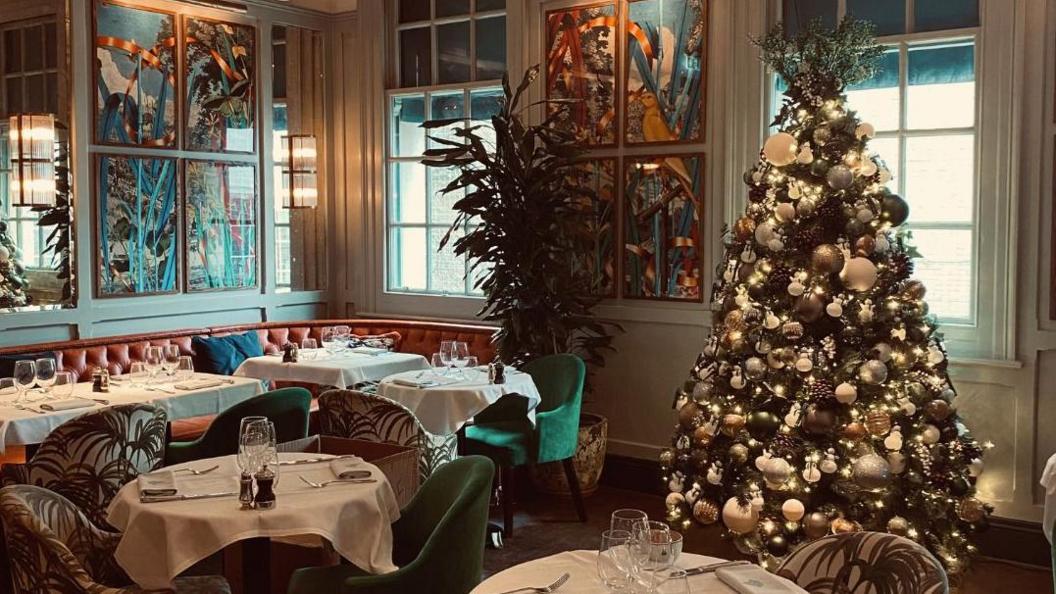 The inside of a restaurant. Table and chairs can be seen alongside a decorated Christmas tree.