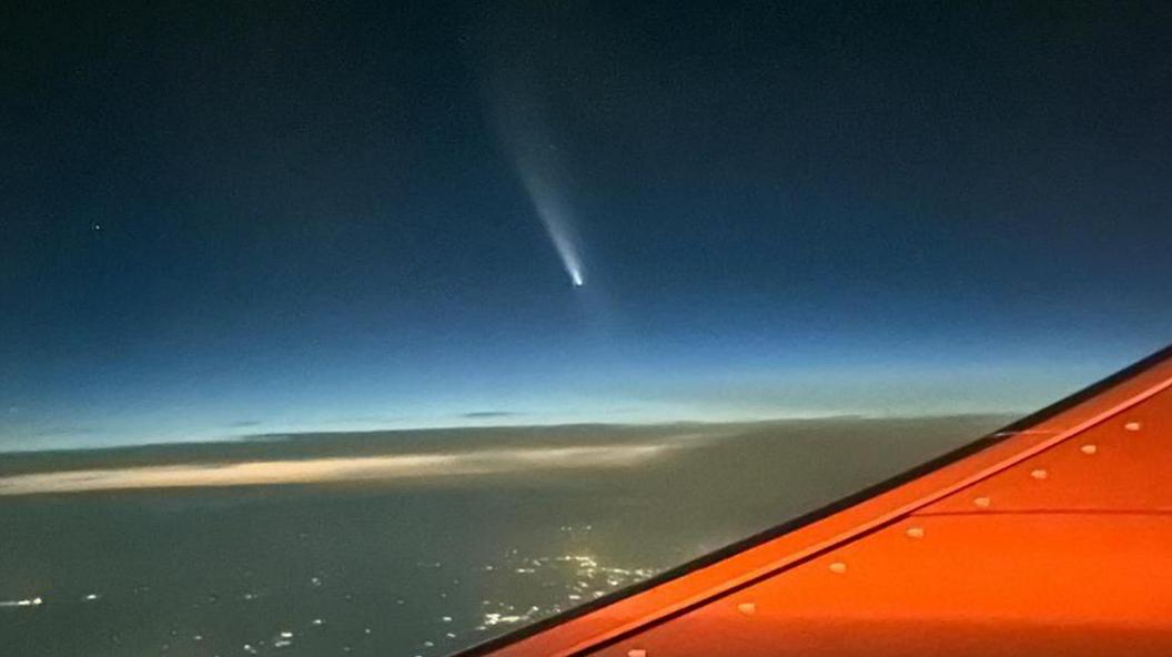 The comet above clouds and city lights. Part of an aircraft's red wing is also visible.