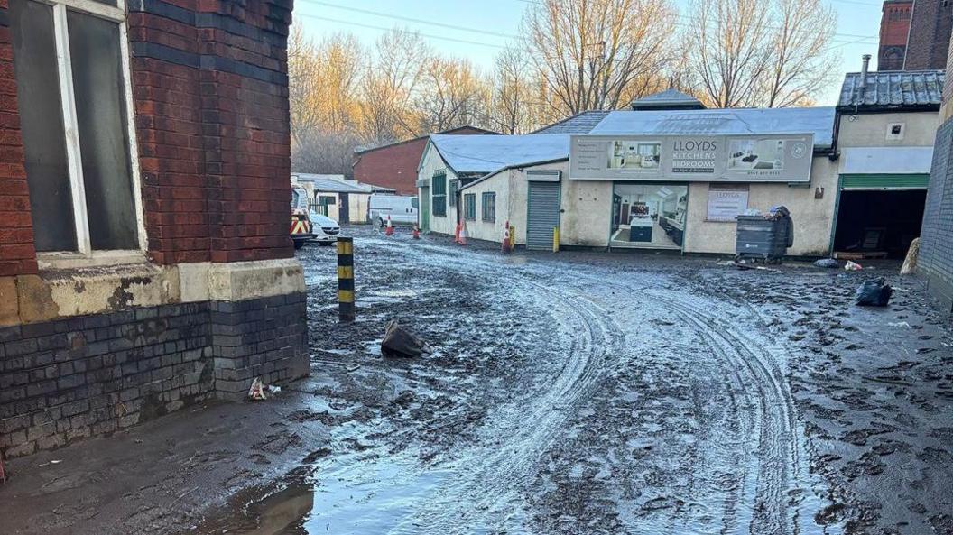 Mud strewn over the ground at Meadow Mill industrial estate. Vehicle tracks and footprints can be seen in the mud, which has been deposited all over the area after heavy flooding. 