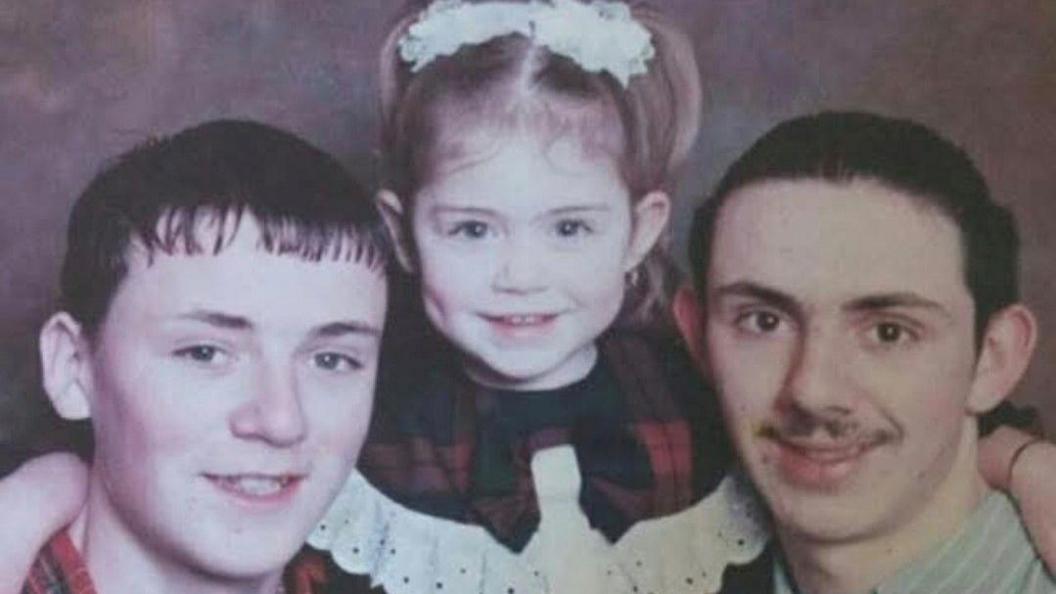 Terry Burns with short brown hair, his sister Danielle with pigtails and wearing a tartan-and-white lace dress and brother Gary with short brown hair and a moustache. All are smiling.