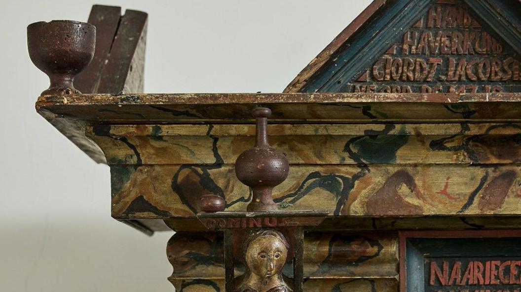 The corner of an ornately carved cupboard, with lettering and what looks like a wooden wine glass carved out of the wood.