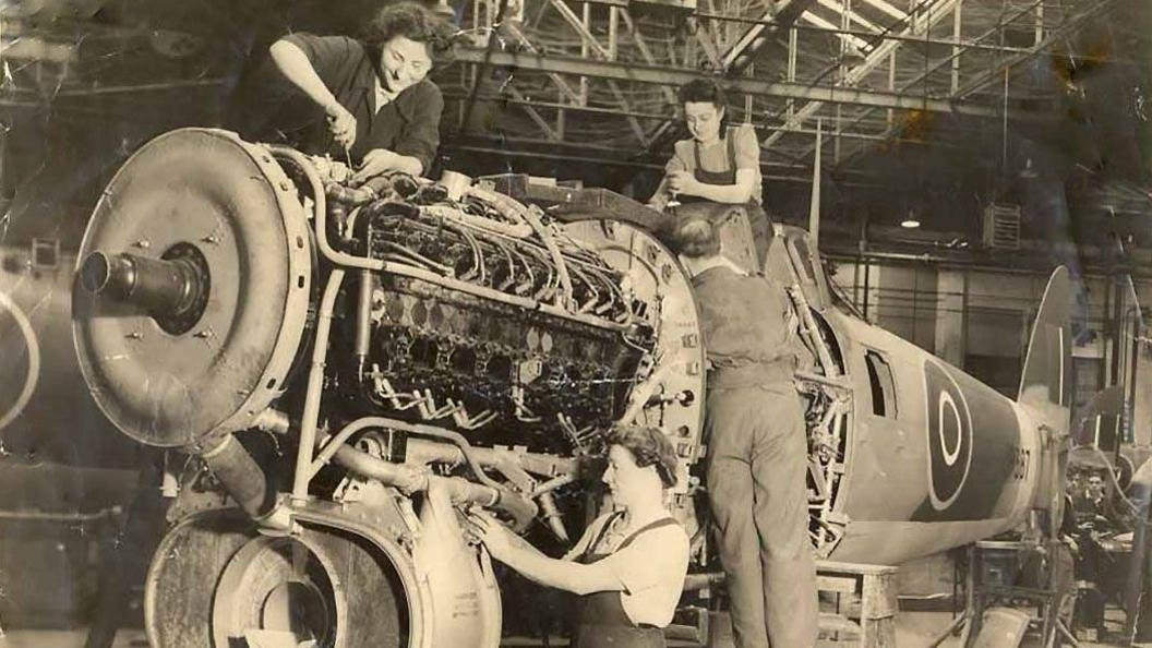 A black-and-white photograph showing a Typhoon aircraft being assembled. Three women are working on parts of the aircraft. A fourth worker, a man, is partly visible with his back to the camera as he fits a component. The factory setting is industrial.