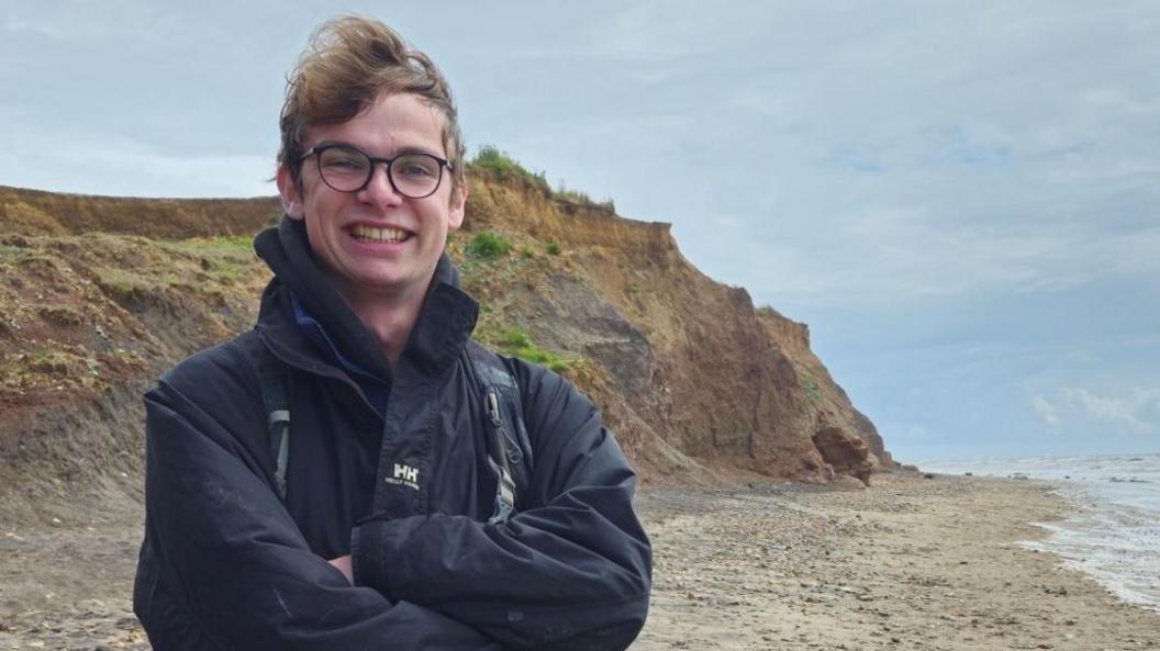 A smiling Joe Thompson standing with his arms folded on a beach. Behind him are some low sloping sand-coloured cliffs. He is wearing a dark-coloured winter jacket and dark-rimmed glasses.