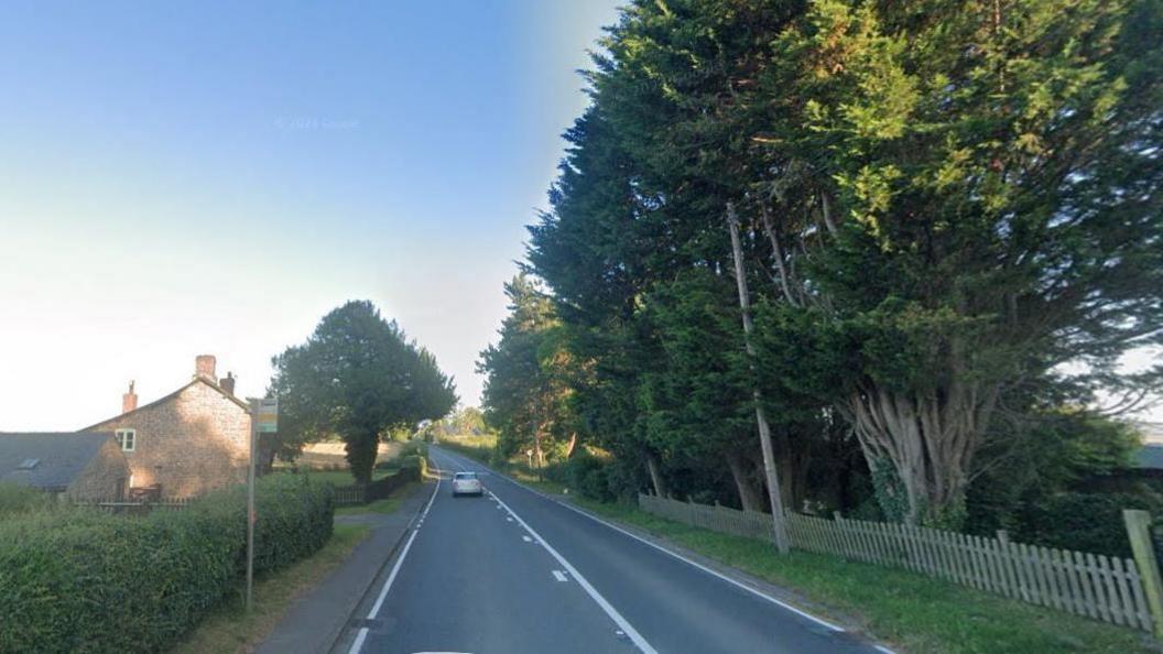 A country road on a sunny day. There are trees behind a fence lining the right side of the road, and there is a bus stop and a large brick building on the left. There is one car driving down the road.