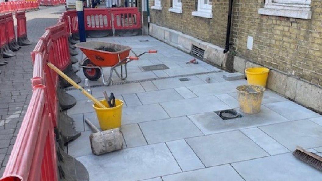 A wheelbarrow and builders items left on the ground for pavement work to be carried out. You can see three buckets, a red wheelbarrow, clean paving and red barriers. There is a brick wall to the right. 