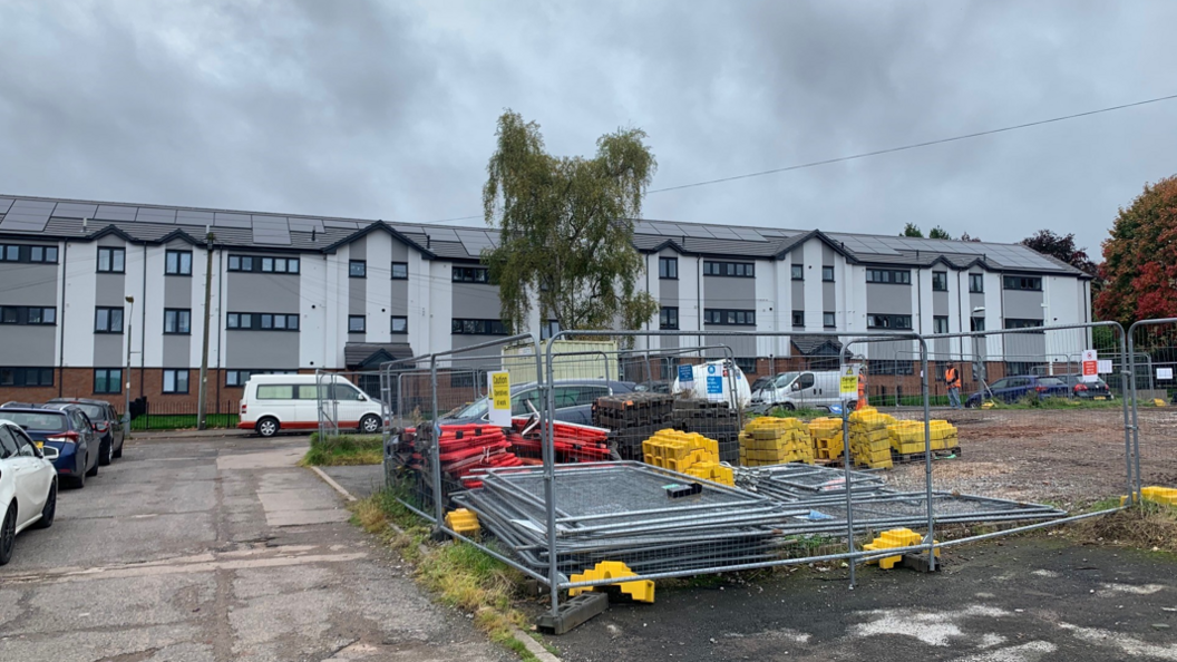 The flats are on several storeys with a grey external design. There are cars parked in front of the building and there is also building work equipment kept inside railings.