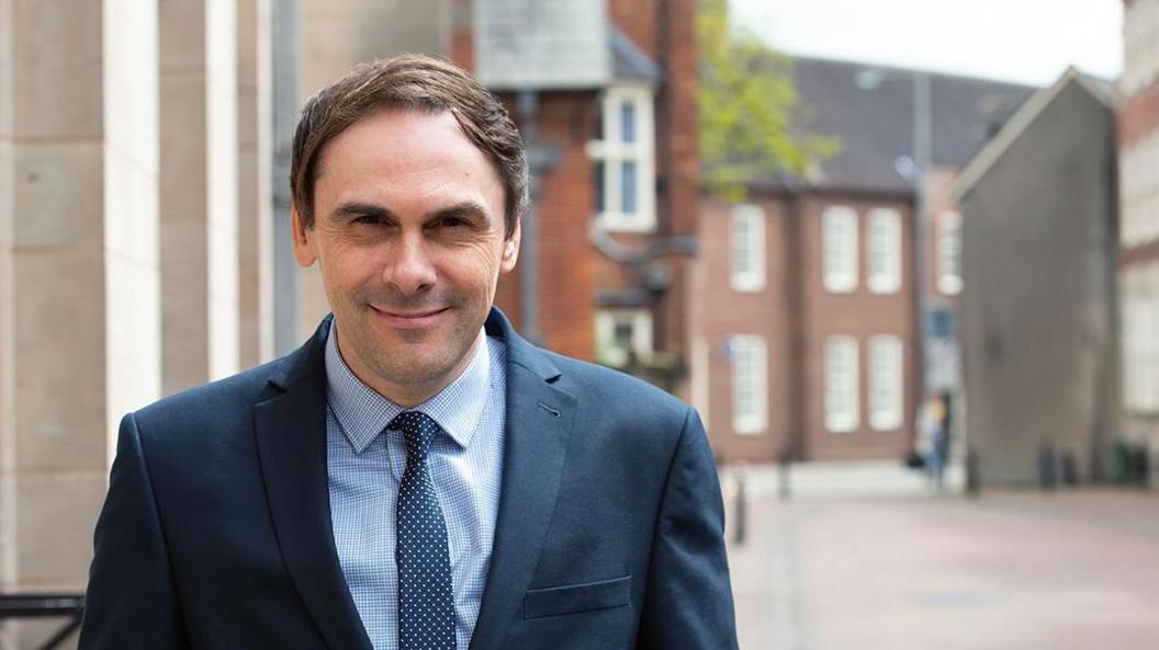 Simon Tagg, a man with brown hair, is wearing a blue suit and spotted navy coloured tie. He is standing in a town centre, with buildings and pedestrian area visible behind him.