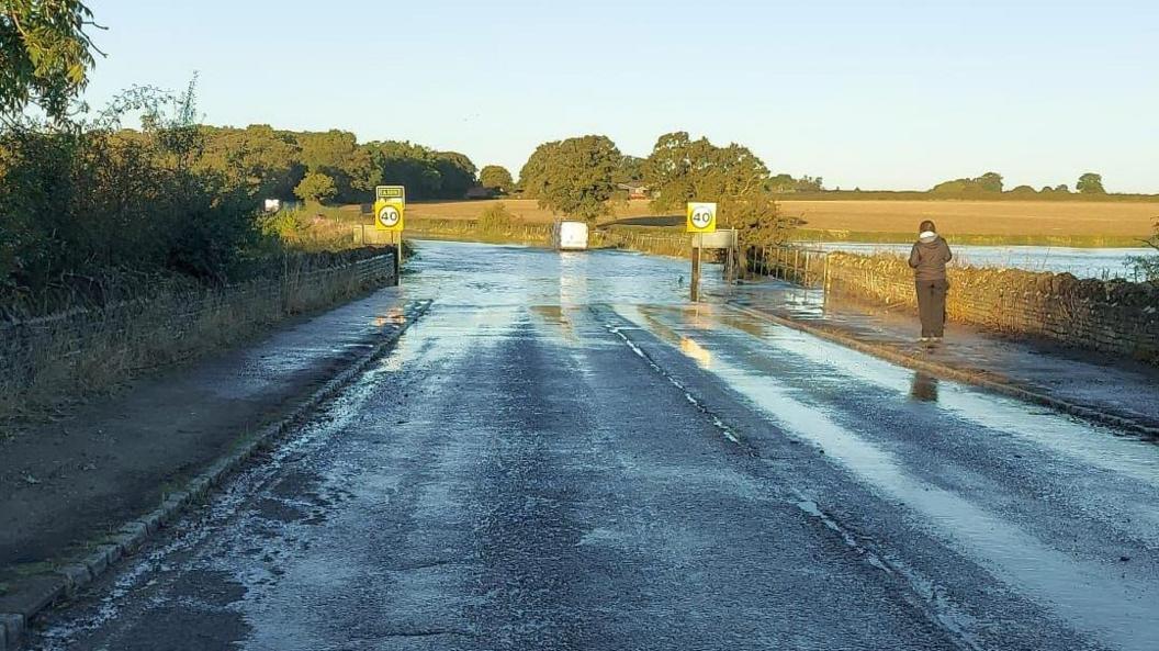 Turvey Bridge in Bedfordshire, it shows a road, a bridge, with lots of water on it, 40mph sings and a person walking towards the bridge